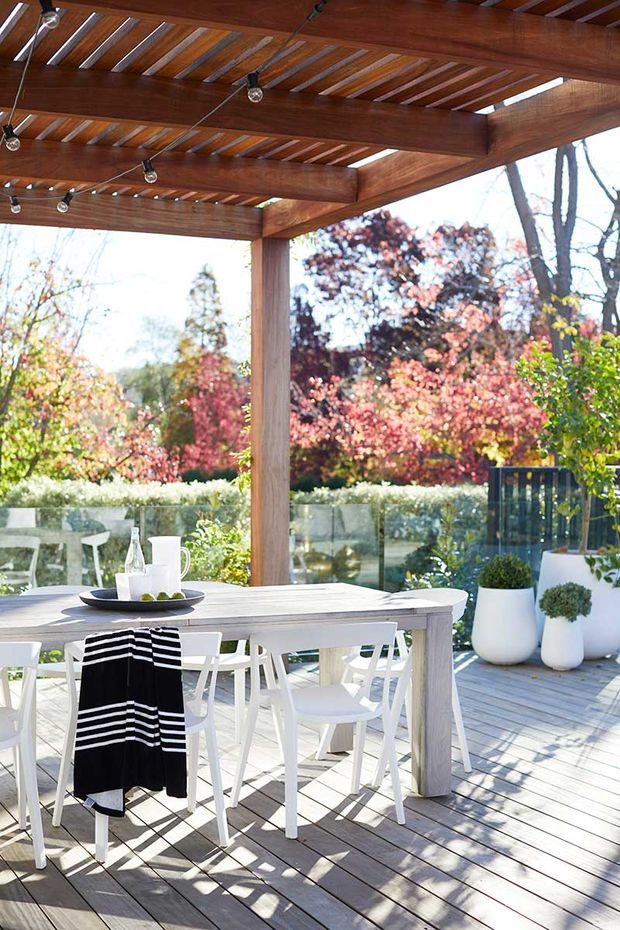 Bright outdoor dining area on a timber deck beneath a substantial timber pergola with exposed beams, slatted roof, and string lighting. A pale timber dining table and white chairs are arranged for entertaining, with tableware and potted plants. Glass balustrading opens to a landscaped garden with autumn foliage.