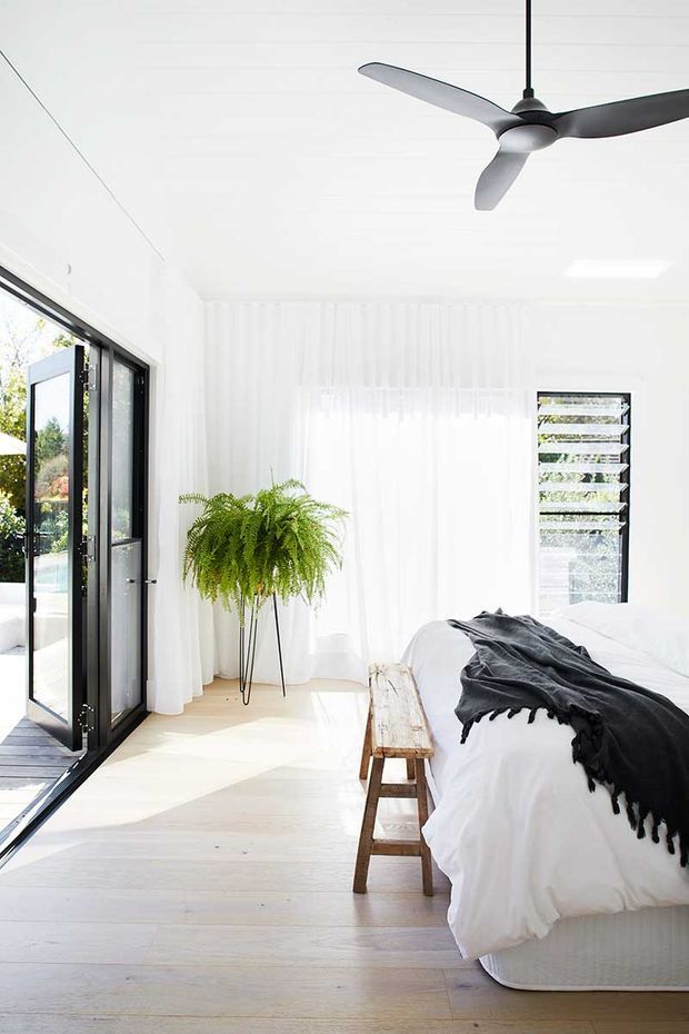 Bright, airy contemporary bedroom with white-painted walls and sheer floor-to-ceiling curtains. A large black-framed glazed door opens to an exterior deck, while a louvred window provides ventilation. The room features pale timber flooring, a white bed with dark charcoal throw, a simple timber bench, a hanging pendant ceiling fan, and a tall fern adding natural greenery.