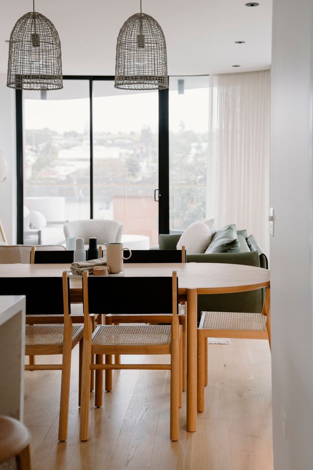 Bright contemporary dining area with a light timber dining table and woven-seat timber chairs, positioned beside a living area. Two oversized woven pendant lights hang above the table, while full-height black-framed glazing and sheer curtains provide abundant natural light. Neutral finishes and muted green upholstered seating create a calm, warm interior.