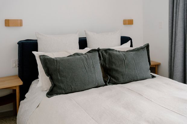 A softly lit bedroom featuring a neatly made bed with white linen bedding and large charcoal-grey cushions, a dark upholstered headboard, simple timber bedside tables, wall-mounted timber reading lights, and full-length grey curtains. The styling is restrained and contemporary with a calm neutral palette.