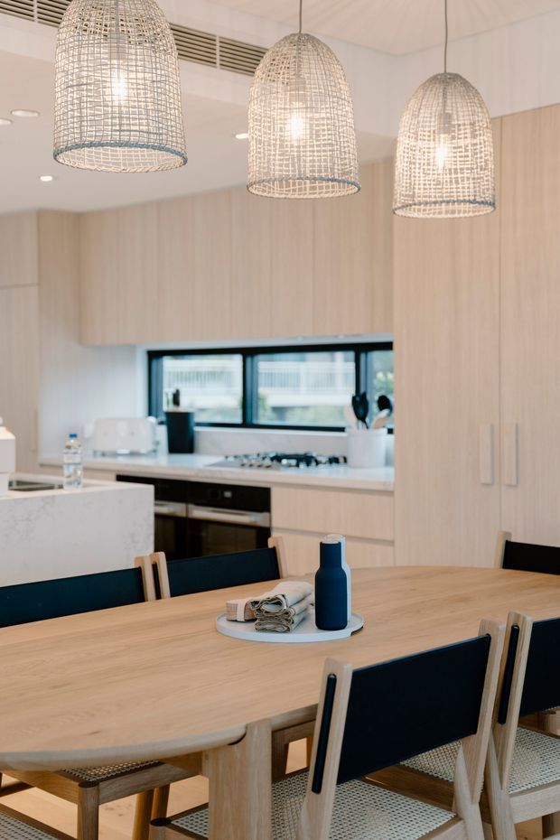 Bright open-plan kitchen and dining area with pale timber cabinetry, a large light-wood dining table, dark blue upholstered dining chairs, white benchtops, and three woven pendant lights. Black-framed windows and integrated appliances add contrast to the warm, contemporary interior.