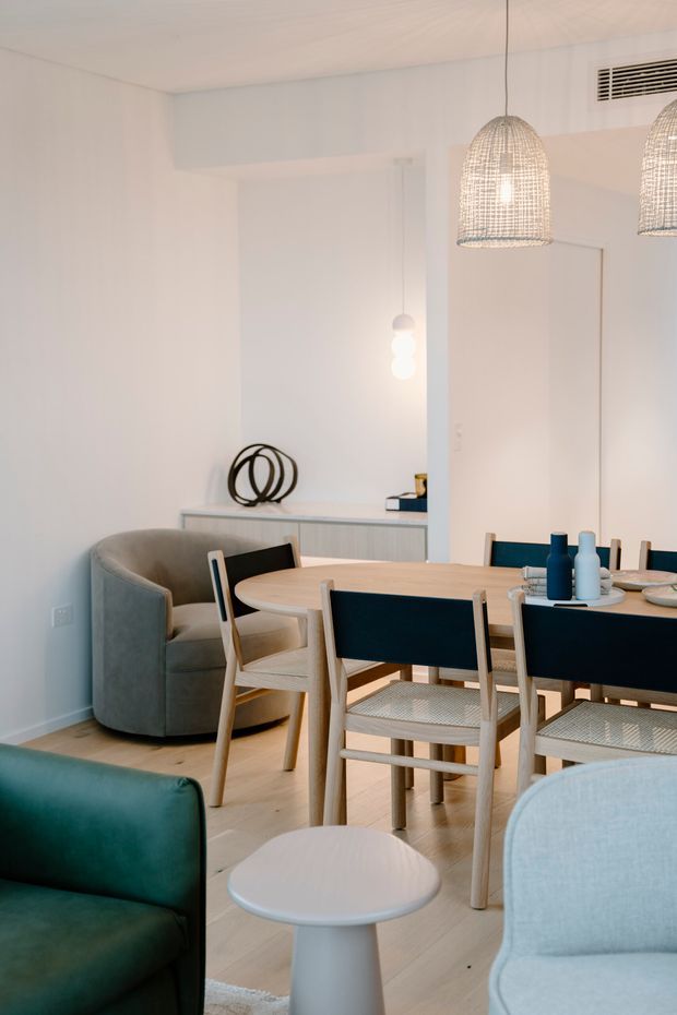 A bright, softly styled open-plan dining and living area with a light timber dining table and chairs, upholstered armchairs, teal seating, a pale wood floor, white walls and ceiling, and woven pendant lights. A slim sideboard with sculptural decor sits against the rear wall.