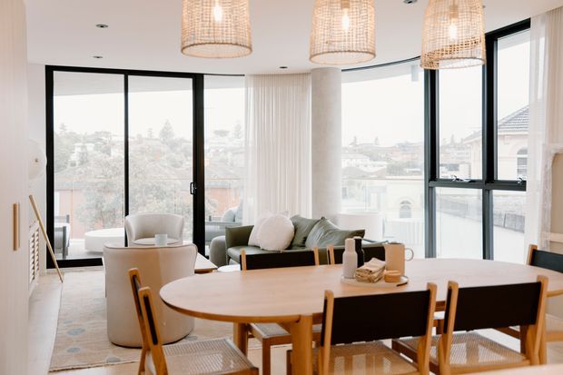 Bright contemporary open-plan living and dining area with floor-to-ceiling black-framed glazing, a large oval timber dining table, lounge seating, pale furnishings, and woven pendant lights. A city or suburban streetscape is visible through the windows.