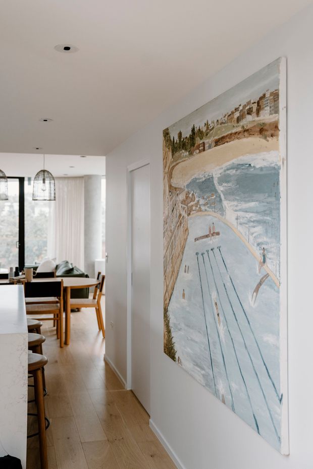A bright, minimalist open-plan residential interior viewed along a hallway beside a kitchen. The foreground features a large framed coastal artwork on a white wall, while the background shows a timber dining table and chairs, pendant lights, pale curtains, and expansive glazing. Light timber flooring and white walls create an airy, warm atmosphere.