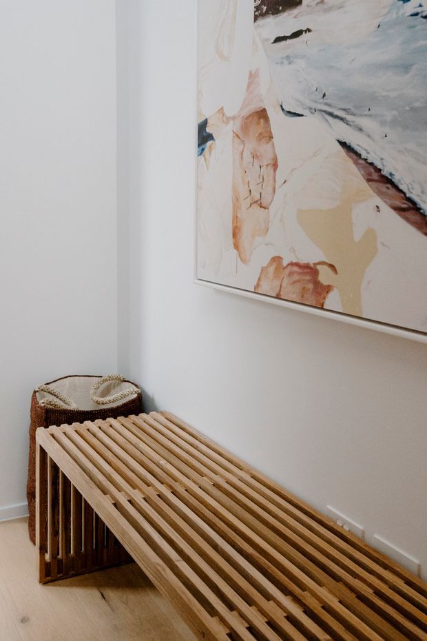 A close-up interior vignette showing a sculptural slatted timber bench or console against a white wall, with a woven basket tucked beside it and a large abstract artwork mounted above. The space has light timber flooring and a restrained, natural contemporary aesthetic.