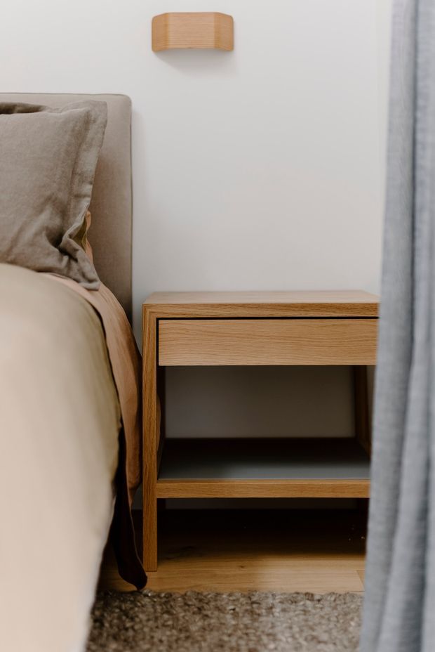 Close-up of a calm, contemporary bedroom corner showing a soft upholstered bed with neutral bedding, a timber bedside table with an open shelf, a matching timber wall light, pale wall, curtain, and warm timber flooring.
