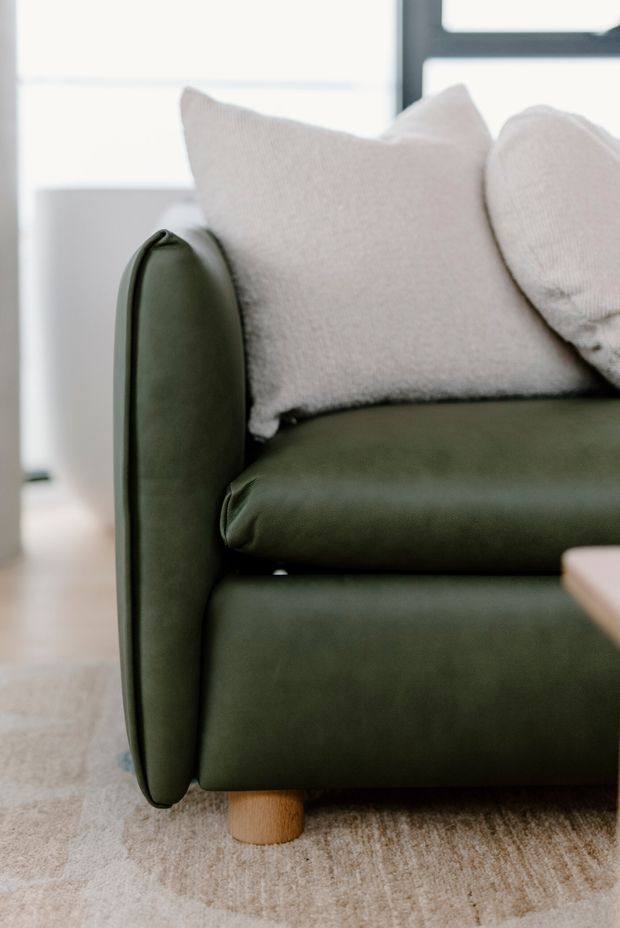 Close-up interior detail of a contemporary sofa with deep olive-green upholstered leather or faux-leather seating and rounded sides, paired with soft off-white textured cushions. A light timber cylindrical leg, pale rug, and bright window provide a calm, refined living-room setting.