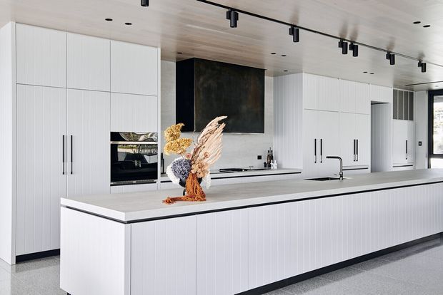 A bright, contemporary galley-style kitchen dominated by a long white island with integrated sink and black tap. Full-height white vertical-panel cabinetry lines the back wall, with built-in ovens, concealed storage and a dark stone or marble-look splashback feature. The ceiling has light timber panelling and black track spotlights, while dark window and door frames add contrast. A dried floral arrangement sits on the island.