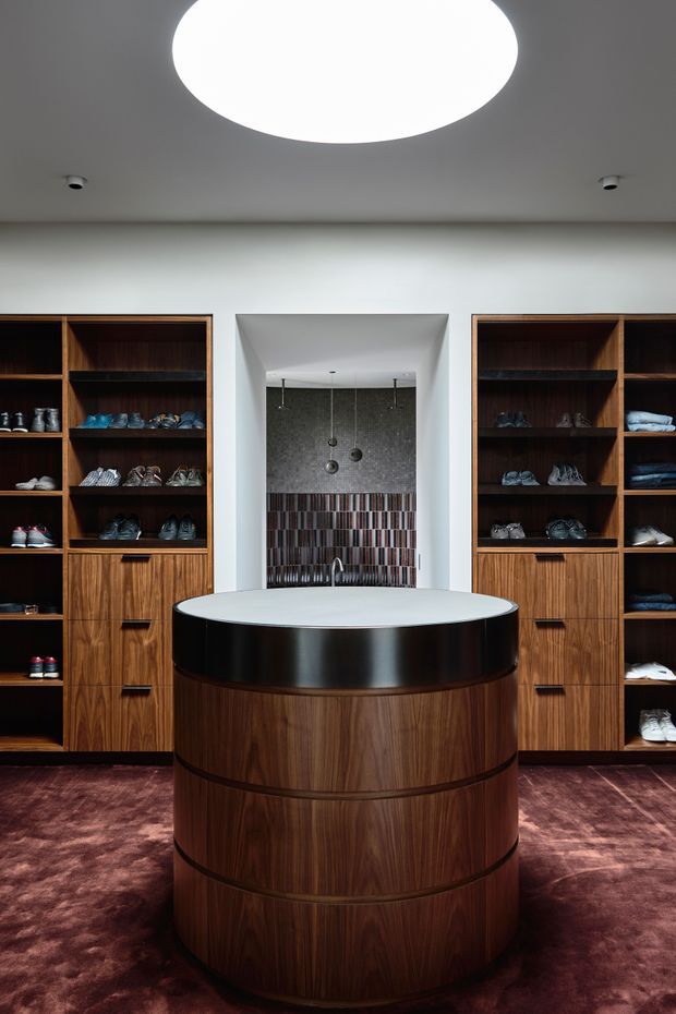 A refined walk-in wardrobe with dark timber built-in shelving displaying shoes and folded clothing on both sides. A curved central island features rich wood veneer, a glossy black band, and a pale stone or solid-surface benchtop. At the rear is a recessed tiled niche with a small basin and pendant lights. The room has a deep burgundy carpet and a large circular ceiling light, creating a luxurious, high-contrast interior.