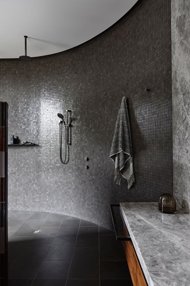A dark, refined bathroom with a curved wall fully clad in small charcoal-grey mosaic tiles. The space features a black walk-in shower with exposed shower fittings, a large-format dark tiled floor, a grey stone vanity benchtop with timber cabinetry, and a rough-textured stone wall at right. A sculptural curved ceiling and minimal black fixtures add a dramatic architectural character.