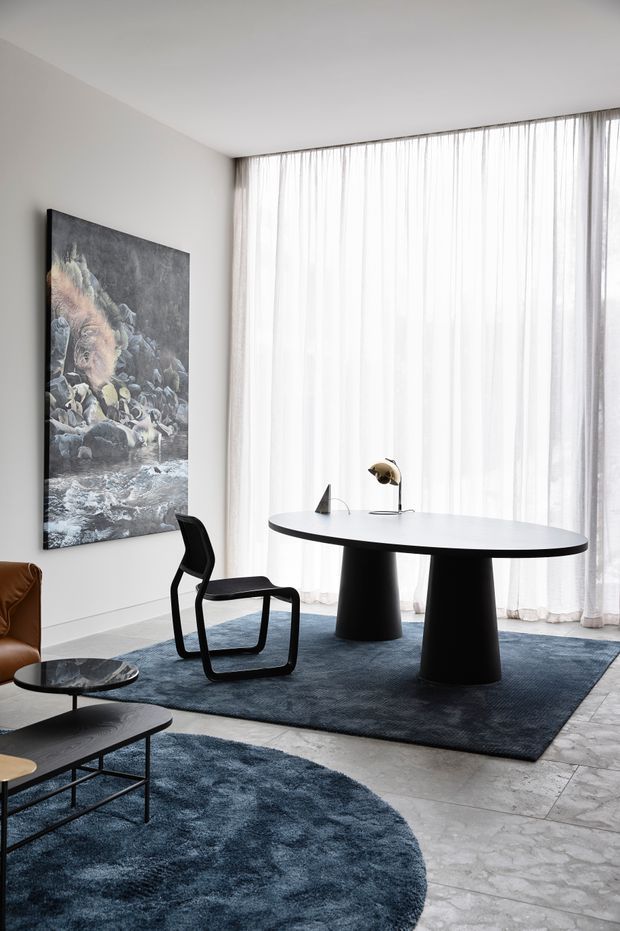 A bright, refined contemporary dining and living area with floor-to-ceiling sheer curtains, a large oval white dining table on sculptural black pedestal bases, a black modern dining chair, dark blue rugs, a leather sofa edge, and a marble-topped coffee table. A large monochrome artwork provides a dramatic focal point against the light walls, while daylight filters through the expansive glazing.