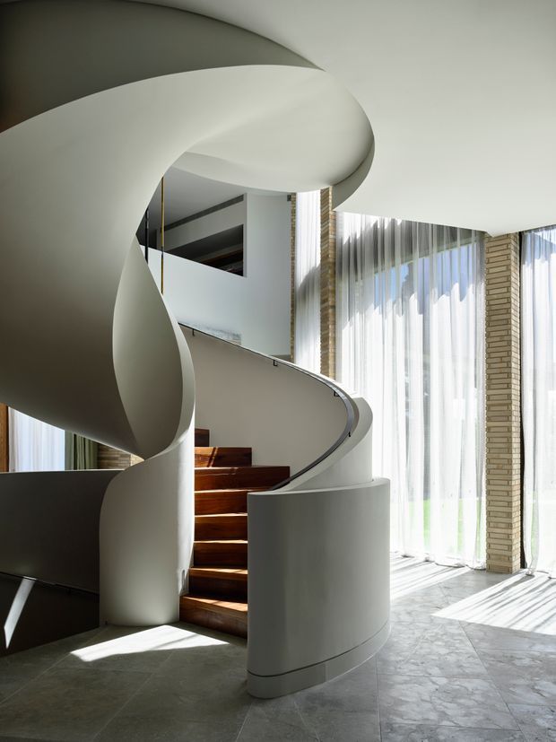 A dramatic sculptural spiral staircase with a smooth white curved balustrade and central form, rising through a bright double-height interior. Warm timber stair treads contrast with pale stone or concrete flooring, while full-height sheer-glazed windows and exposed light brick columns flood the space with natural light.
