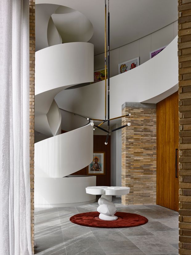 A dramatic multi-level interior foyer featuring a sculptural white spiral staircase with curved balustrades, a tall central void, and a contemporary suspended branching light fixture. The space has grey stone tile flooring, exposed warm brick walls and columns, a timber pivot door, framed artwork, and a sculptural white console table on a round red rug. Sheer curtains frame the view from the side.