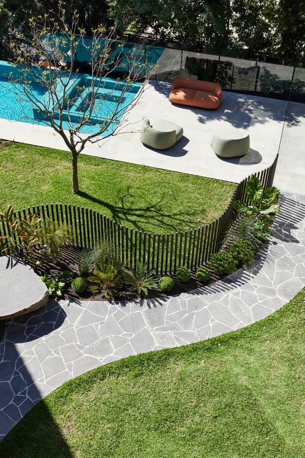 A bright landscaped residential backyard viewed from above, featuring a vivid turquoise swimming pool, a pale paved pool terrace with sculptural lounge seating, manicured lawn, a curved dark vertical-slat fence, tropical planting, and an irregular dark stone garden path. The space has a contemporary, carefully designed outdoor entertaining and pool setting with strong geometric curves and contrasting textures.