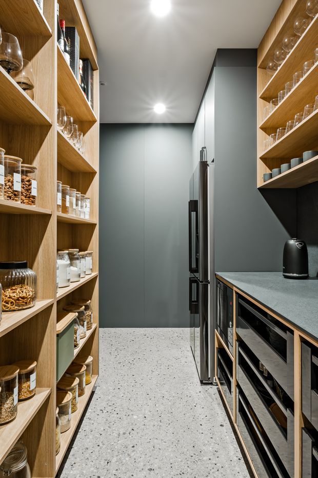 A narrow, highly organized walk-in pantry with full-height open timber shelving on both sides, stocked with jars, glassware, cups, and food containers. A dark grey kitchen wall with integrated stainless-steel refrigeration and a long grey stone benchtop occupies the rear and right side. The floor is finished in light terrazzo with dark aggregate, and recessed ceiling lights provide bright, even illumination. The design combines warm timber joinery with a dark contemporary palette.