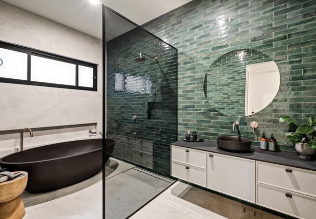 A contemporary bathroom featuring a freestanding matte black bathtub, walk-in glass shower enclosure, dark green brick-pattern wall tiles, a floating white vanity with dark benchtop, round vessel basin, and large circular mirror. The space has neutral plastered walls, concrete-look flooring, black-framed glazing, and a clean, refined palette.