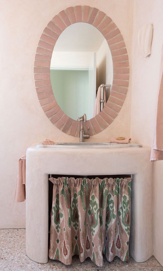 A compact, softly styled bathroom vanity with a rounded plastered basin surround, wall-mounted oval mirror framed in pink brick tiles, aged metal faucet, and patterned fabric curtain concealing under-sink storage. Pale blush walls, terrazzo flooring, and muted green, pink, and cream textiles create a warm, decorative interior.