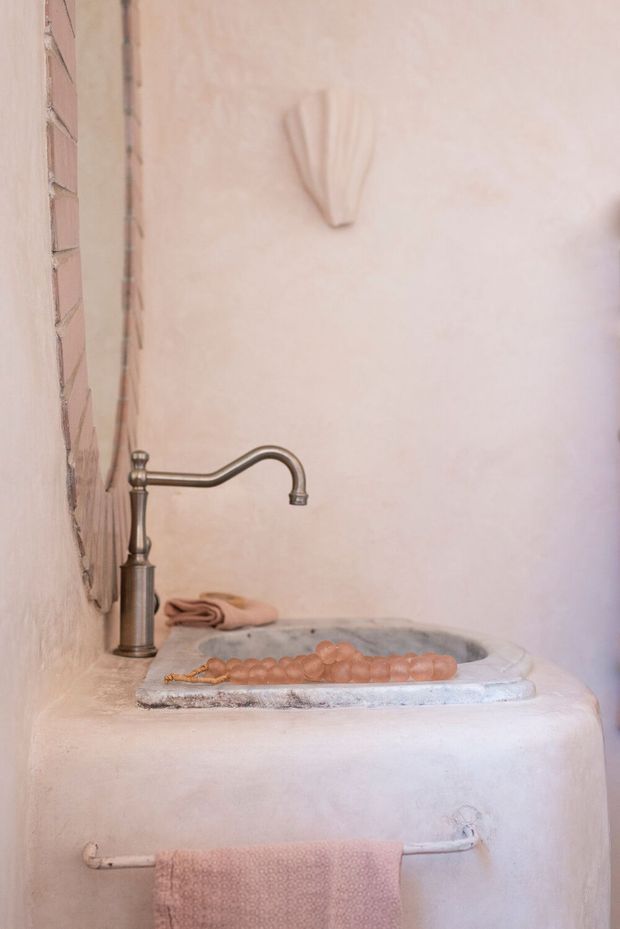Close-up of a handcrafted bathroom vanity with a raised, integrated basin, aged bronze-style curved tapware, pale plaster walls, and a dusty pink towel. The rustic, sculptural basin surround and soft blush palette create a warm, artisanal character.