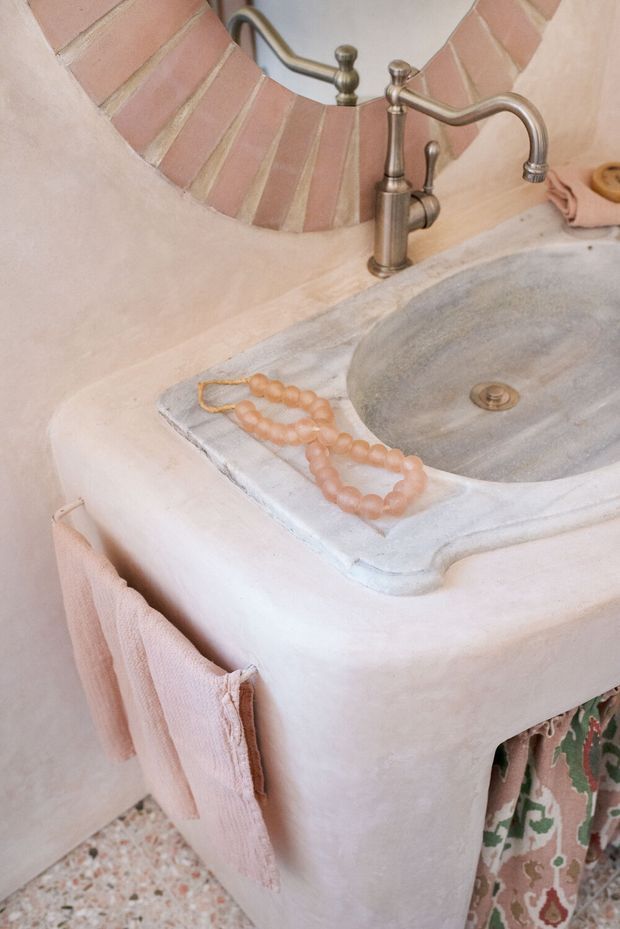 Close-up of a softly styled bathroom vanity with a rounded plaster or stucco base, pale stone or concrete countertop, integrated oval basin, and aged brass bridge-style faucet. A curved mirror with a terracotta-toned tiled surround is mounted above the vanity. Blush towels and a beaded accessory add warm decorative details, while terrazzo flooring is visible below.