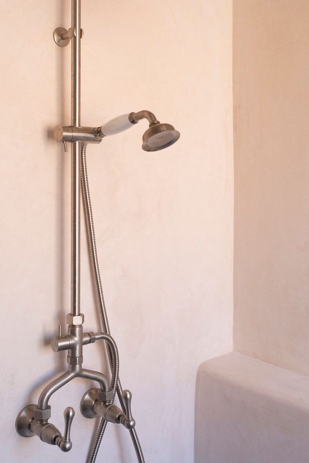 Close-up of a compact shower and bath fixture installation in a pale plastered bathroom. The exposed plumbing and shower rail have a brushed metallic finish, with a handheld shower hose, controls, and a rounded built-in bathtub edge visible at the lower right.