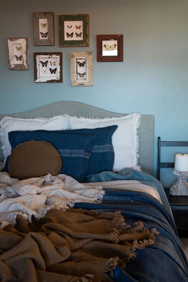A cozy bedroom vignette featuring a bed with a softly curved upholstered headboard, layered navy, white, blue and brown bedding, and fringed textured throws. A small dark bedside table with a lamp sits to the right, while a collection of framed butterfly artworks decorates the muted blue wall above the bed.