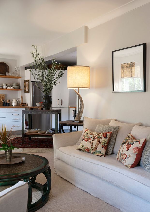 A warm, refined open-plan living and kitchen interior. A light upholstered sofa with patterned cushions fills the foreground, facing a round dark wood and glass coffee table. Behind it, a compact kitchen features white cabinetry, open timber shelving, dark benchtops, and stainless-steel appliances. A sculptural floor lamp, framed artwork, indoor greenery, and a dark patterned rug add character. The room has white walls and ceiling with simple traditional trims and abundant natural light.