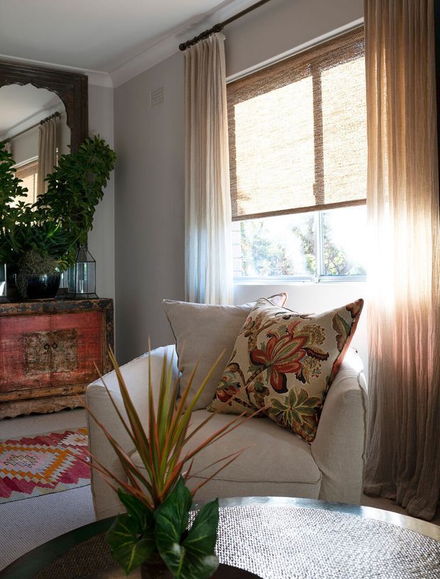A warmly lit, traditionally styled sitting area with a cream upholstered armchair, patterned floral cushion, leafy indoor plant, antique carved wooden chest, decorative mirror, patterned rug, and a large window with woven roller blinds and floor-length curtains. The palette is neutral and earthy with colourful textile accents.