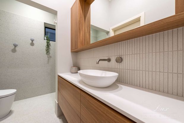 Bright contemporary bathroom featuring a floating timber vanity with a white countertop, round vessel basin, brushed metal wall-mounted tapware, and a large timber-framed mirrored cabinet. The shower area has light textured wall and floor finishes, with a partially visible wall-hung toilet and a small high window.