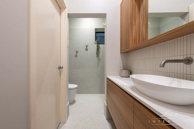A contemporary bathroom with a floating timber vanity, white vessel basin, wall-mounted brushed metal tapware, vertical light tile splashback, large framed mirror, and a separate toilet and shower area. The space has a warm neutral palette and clean built-in joinery.