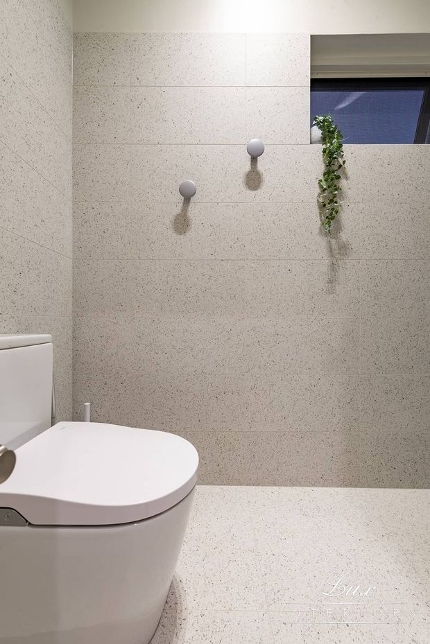 A compact contemporary bathroom featuring a white toilet, pale speckled large-format wall and floor tiles, minimalist spherical wall hooks, and a narrow high window with a trailing indoor plant. The space has a clean, restrained neutral palette and integrated tiled surfaces.