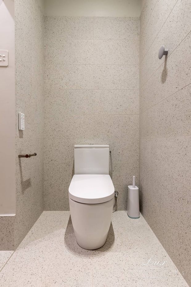 Compact modern toilet room clad in light speckled stone-look tiles across the walls and floor. A white wall-hung-style toilet, minimalist flush controls, toilet paper holder, and toilet brush create a clean, understated fit-out.
