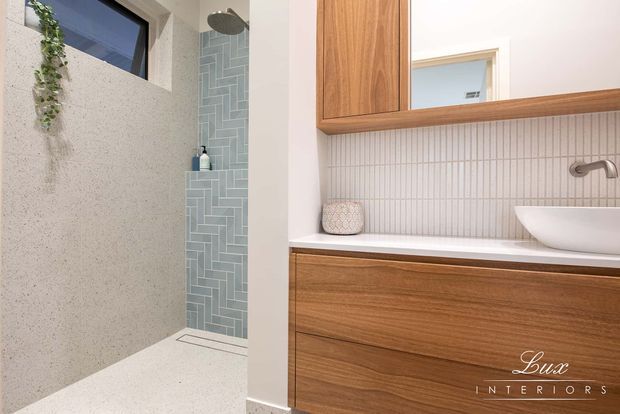 A contemporary bathroom featuring a walk-in shower with pale blue-green vertical herringbone tiles, a textured light terrazzo-look wall and floor, and a timber vanity with integrated drawers. The vanity has a white benchtop, fluted light tile splashback, round vessel basin, brushed metal wall-mounted tapware, mirrored cabinet, and a small high window with trailing greenery.
