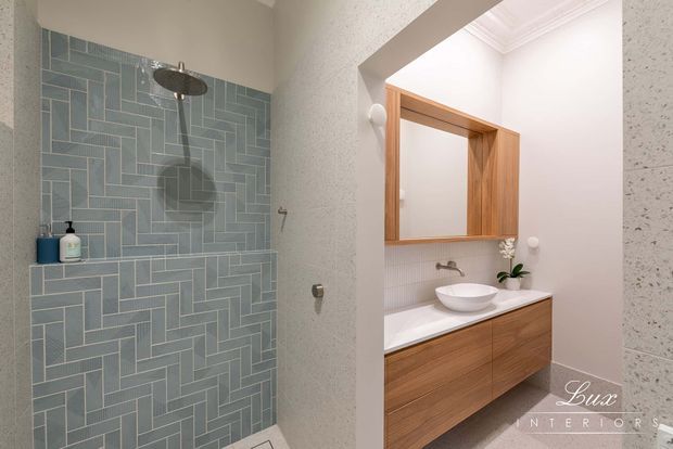 Bright contemporary bathroom featuring a walk-in shower with a blue-grey geometric tiled feature wall, wall-mounted shower fittings, and a floating timber vanity with a white vessel basin. A recessed timber-framed mirrored cabinet, white benchtop, small plant, and light textured walls complete the refined fit-out.