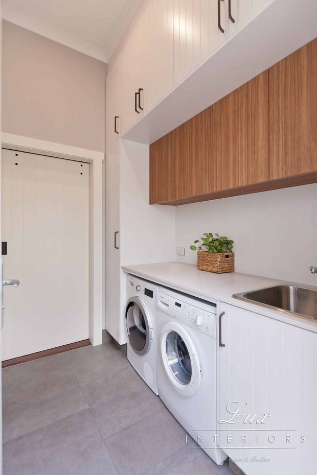 Bright, compact laundry room with front-loading washing machine and dryer beneath a pale benchtop, white lower cabinetry, white vertical-panel overhead cabinetry, and a warm timber wall-mounted storage unit. A stainless-steel sink is integrated at the right, with grey tiled flooring, neutral painted walls, and a small potted plant adding a natural accent.