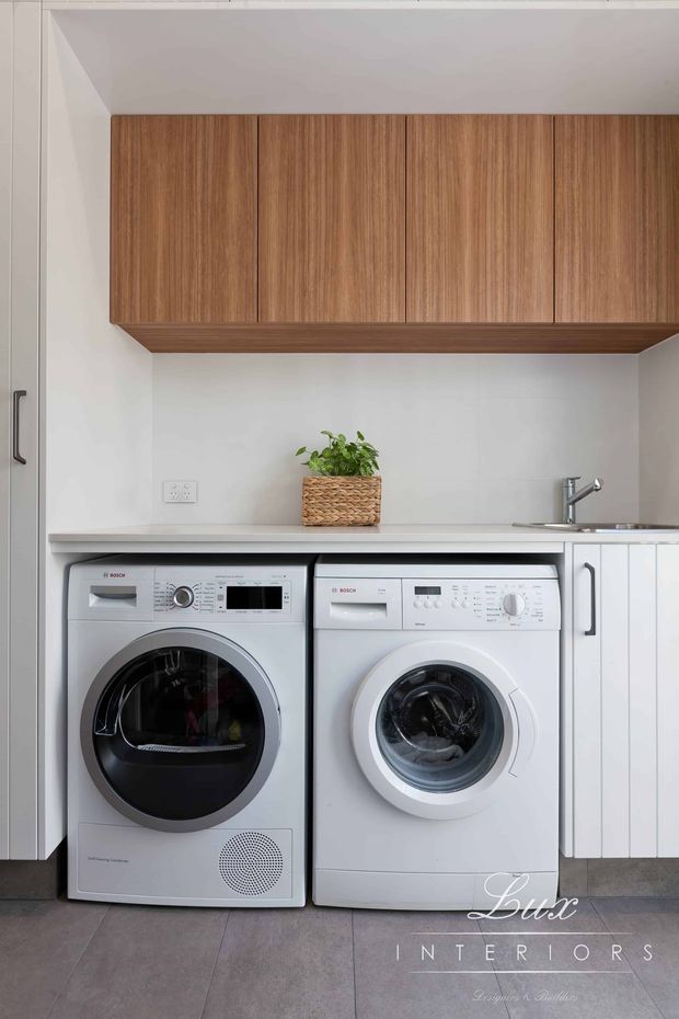 Compact, contemporary laundry room with stacked? side-by-side front-loading Bosch washer and dryer beneath a white benchtop. Warm timber overhead cabinetry spans the wall, with white lower cabinetry, a stainless tap and sink area, a woven planter, and large grey floor tiles.