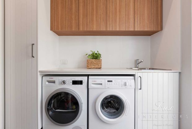 Compact, neatly fitted laundry room with side-by-side front-loading washing machines beneath a white benchtop, integrated white vertical-panel cabinetry, a stainless-steel sink and tap, and warm timber overhead cabinets. The space has a clean, contemporary built-in design with a small woven planter adding natural texture.