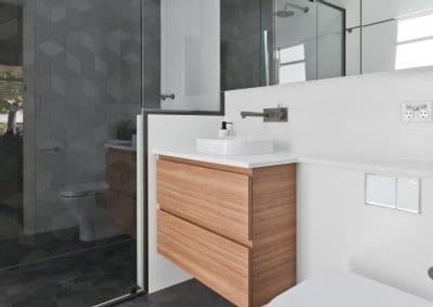 A modern bathroom featuring a floating light-wood vanity with two drawers, a white countertop basin, large wall mirror, frameless glass shower enclosure, white wall surfaces, and dark charcoal floor and shower tiling. A concealed-cistern toilet is partially visible at the right.