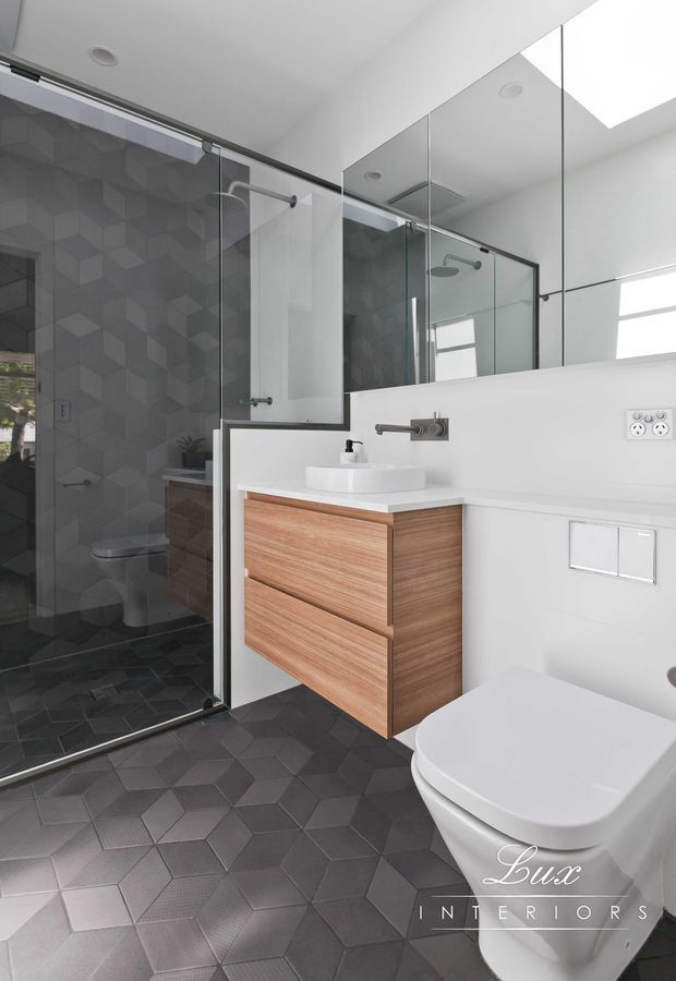 Contemporary bathroom featuring a frameless glass walk-in shower lined with dark geometric cube-pattern tiles, matching dark geometric floor tiles, a floating timber vanity with a white vessel basin, wall-mounted tapware, mirrored cabinetry, and a white wall-hung toilet. The room is bright and clean with strong black-and-white contrast and warm timber accents.