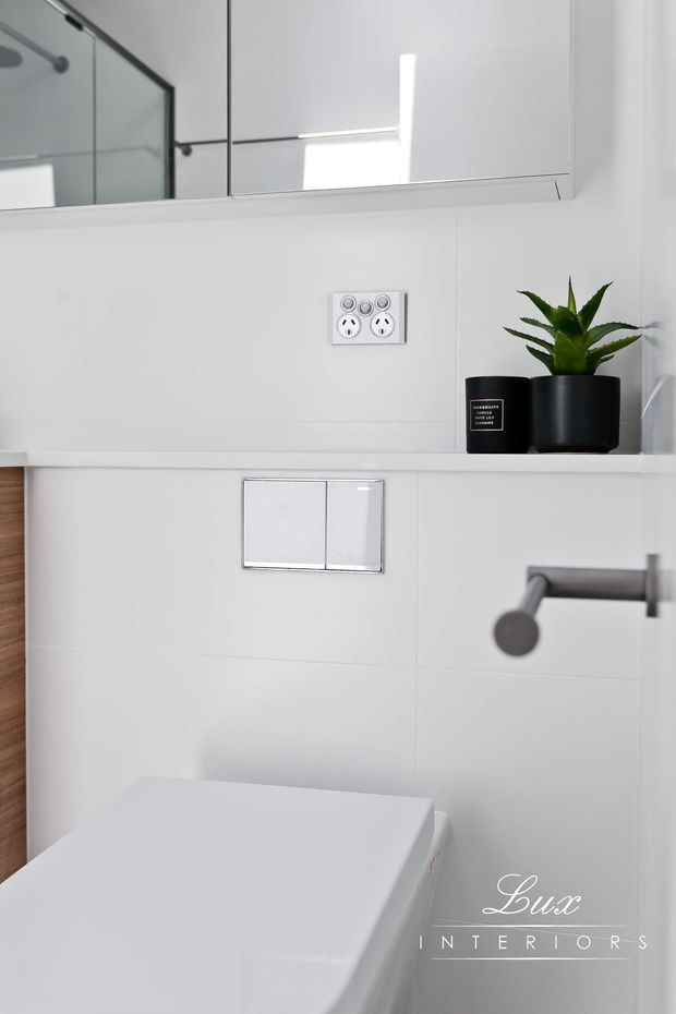 A compact, bright contemporary bathroom showing a wall-hung toilet with dual-flush plate, concealed cistern, wall-mounted brushed-metal tap, mirrored wall cabinet, white tiled walls, and a small black planter on a narrow ledge. The scheme is predominantly white with dark metal accents and clean minimalist detailing.