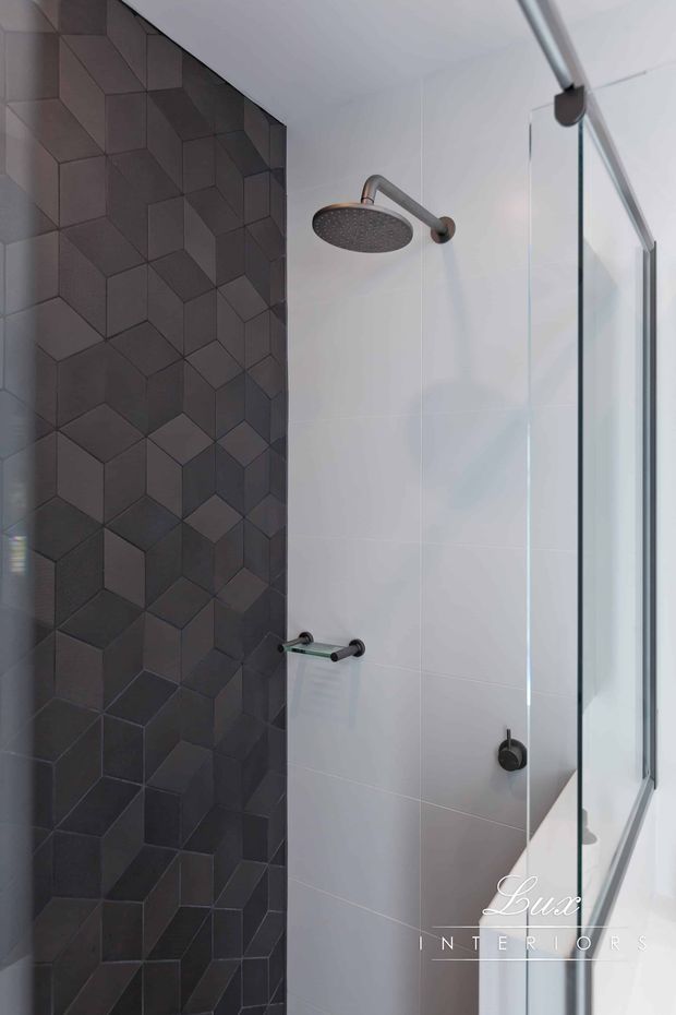 Close-up view of a contemporary shower enclosure with a frameless glass screen, large round showerhead, dark brushed-metal fixtures, and contrasting wall finishes. One wall features dark three-dimensional geometric cube-pattern tiles, while the remaining walls are clad in large pale tiles. The space is clean, refined, and monochromatic.