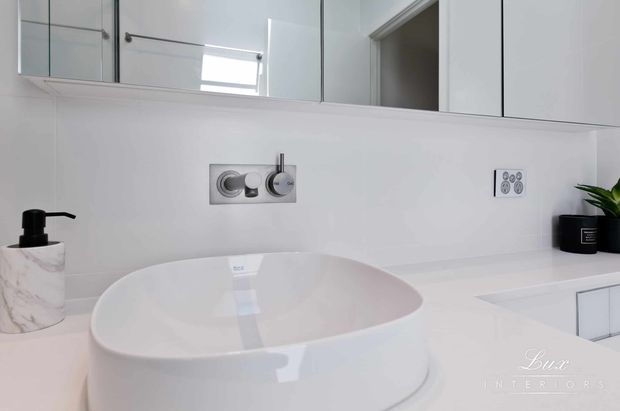 Close-up of a bright, contemporary bathroom vanity with a large white vessel basin on a white benchtop, wall-mounted chrome tapware, mirrored wall cabinetry, and integrated white storage. A soap dispenser and small plant provide minimal accents.