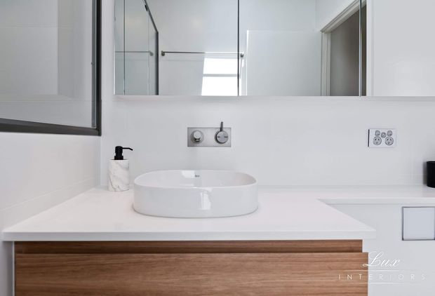 Contemporary bathroom vanity with a white vessel basin set on a white benchtop, wall-mounted chrome tapware and flush controls, a large mirror, and warm timber-fronted cabinetry. The space is clean, minimal, and predominantly white with natural wood accents.
