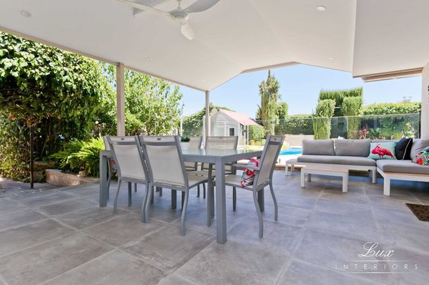 Bright covered outdoor entertaining area with a dining table and chairs, outdoor lounge seating, large-format grey tiled flooring, and a ceiling fan. The space opens onto a landscaped garden and swimming pool, with a clean contemporary resort-like feel.