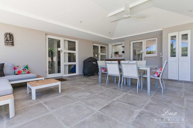 Covered outdoor entertaining area with a dining table and chairs, lounge seating, tiled flooring, pale rendered walls, multiple glazed doors and windows, and a white ceiling fan. The space feels bright, relaxed and connected to the home, with a barbecue visible near the glazing.