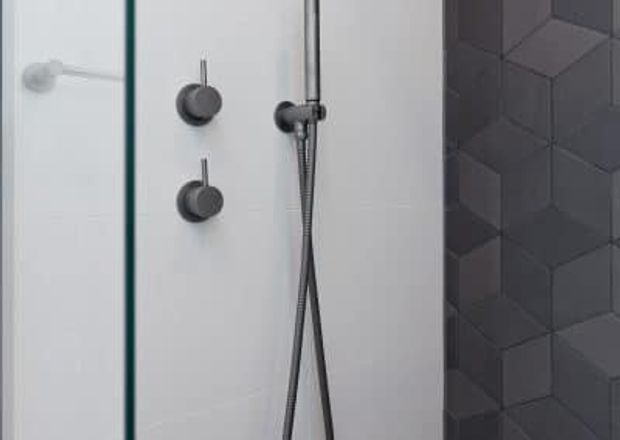 Close-up of a contemporary shower enclosure with clear glass, white wall surfaces, dark brushed-metal shower controls and hand shower, and a dark geometric three-dimensional tiled feature wall.