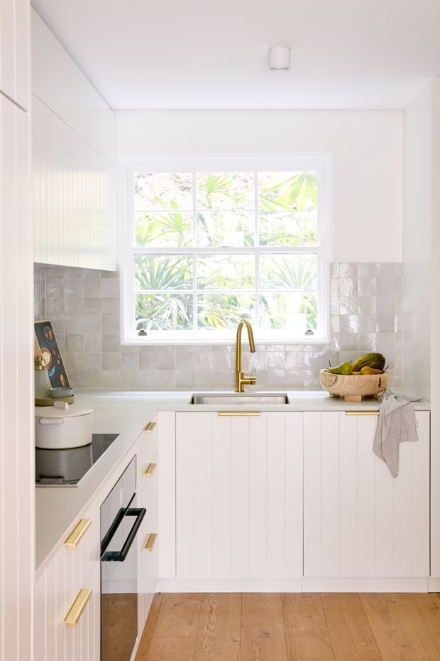 Bright compact kitchen with white vertical-panel cabinetry, brass hardware and tapware, a pale glossy tiled splashback, integrated cooktop and oven, stainless sink, and warm timber flooring. A large divided window overlooks leafy greenery, bringing abundant natural light into the space.