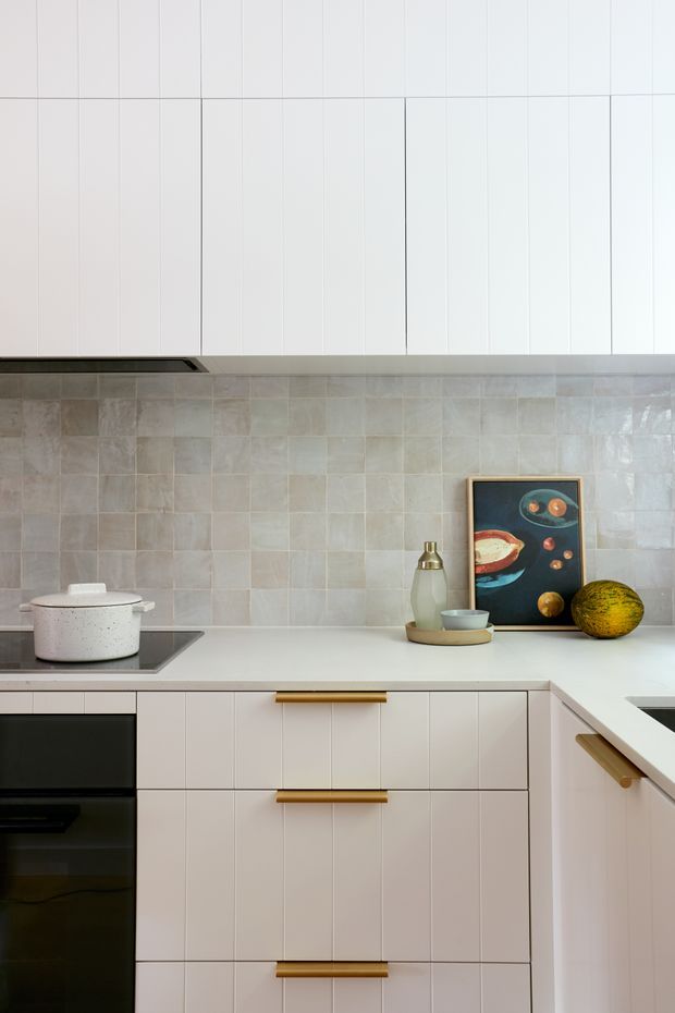 A close-up view of a contemporary kitchen featuring white vertically panelled cabinetry with warm timber handles, a pale stone or solid-surface benchtop, and a light grey-beige square tiled splashback. A black induction cooktop and integrated black oven are visible, with minimal styling including a ceramic pot, vessels, framed artwork and fruit.