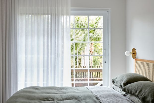 Bright, serene bedroom with a softly made bed in muted grey-green bedding, a woven cane headboard, sheer white curtains, and a brass-and-white wall sconce. A glazed French door opens to a timber balcony or deck with lush palm vegetation beyond.