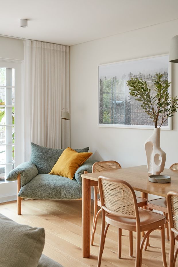 A bright, refined open-plan living and dining area with pale timber flooring, a soft green upholstered armchair, and a light timber dining table surrounded by cane-backed chairs. Floor-to-ceiling curtains frame glazed doors, while a large framed artwork, sculptural ceramic vase, and warm yellow cushion add understated colour and texture. The room has a calm, contemporary natural aesthetic with abundant daylight.