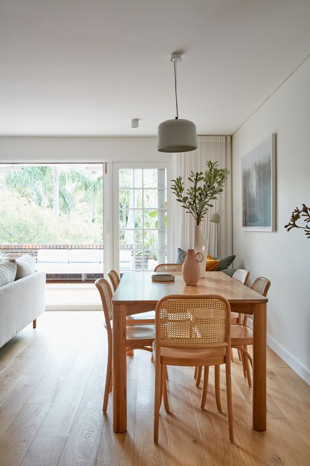 Bright open-plan dining area with a light timber dining table and woven cane chairs, opening into a lounge space and leafy outdoor view. The room features pale timber flooring, white walls and ceiling, a large grey pendant light, vertical wall panelling, framed artwork, and restrained natural-toned styling.
